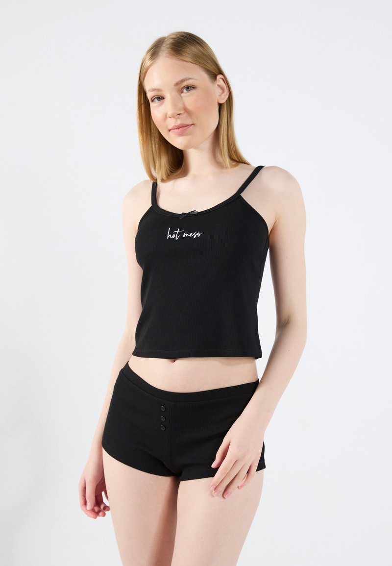 Brassière courte noire côtelée avec des bretelles ajustables, ornée d'un texte brodé blanc "hot mess", associée à un short noir taille haute avec détails de boutons.