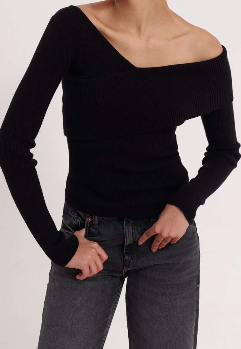 Personne portant un pull noir côtelé à épaules dénudées et un jean gris foncé, ajustant la taille devant avec les mains.