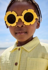 Rumene sončna očala z rožnatim vzorcem in velikimi črnimi lečami; rumena teksturirana srajca z gumbi. Približek obraza in zgornjega dela telesa otroka.