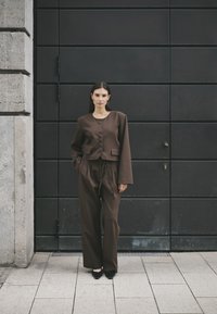 Brun cropped jakke med knapper, parret med højtaljede, brede bukser. Glat tekstur, minimalistisk design, sat mod en mørk baggrund.