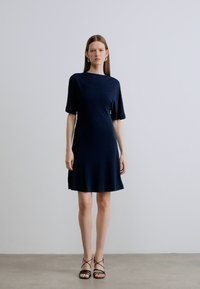 Rochie bleumarin cu mâneci scurte, croială lejeră, tiv evazat și material fin. Asortată cu sandale negre cu barete.