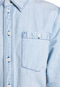 Ljusblå denimskjorta med spetsig krage, bröstficka och vita knappar med kontrasterande sömmar längs sömmarna.