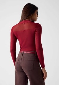 Czerwony prążkowany top z długim rękawem, z przezroczystą górną częścią i ozdobnymi ćwiekami na rękawach, w połączeniu z brązowymi spodniami z wysokim stanem i dopasowanym paskiem.