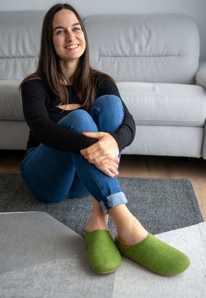 Chaussons en feutre vert avec un design à enfiler et des orteils arrondis, reposant sur un tapis texturé gris. Teintes naturelles sobres et élégantes.
