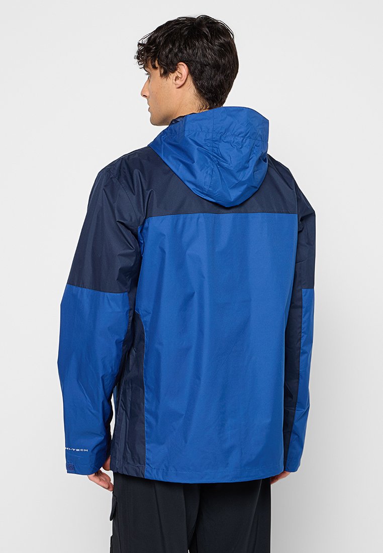 Homme portant une veste imperméable bleu et marine avec capuche, vu de dos, se tenant contre un fond clair.