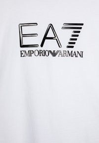 Balta medvilninė marškinėliai su dideliu juodu atspaudu 'EA' su stilizuotomis linijomis, o žemiau - 'EMPORIO ARMANI' tekstūruotu šriftu.