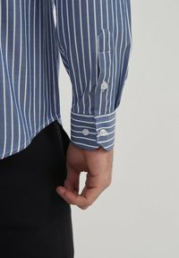 Chemise rayée bleue et blanche avec des poignets boutonnés et un revers contrastant, montrée avec une main tenant l'ourlet.