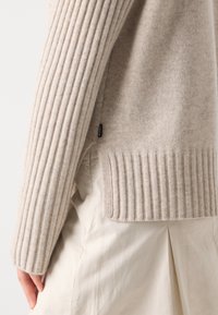 Beige strikket genser med ribbestrikkede ermer og kant, laget av mykt stoff med en subtil tekstur, supplert med en sideløsning og merkeflagg.