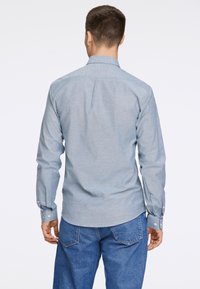 Camisa azul clara de botões, com mangas longas e colarinho padrão, apresentando um tecido texturizado e punhos com botão, usada com jeans azuis.