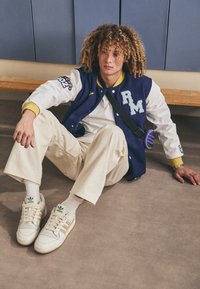 Młoda osoba z kręconymi włosami siedząca na podłodze, ubrana w niebiesko-białą kurtkę varsity, beżowe spodnie, białe adidasy i małą torebkę na ramię.