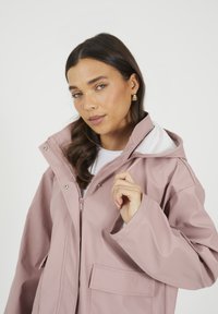 Veste imperméable rose avec un col haut, fermeture éclair et poches latérales. Possède une doublure blanche et une texture lisse.