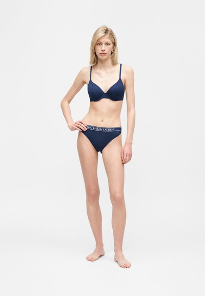 Ξανθιά γυναίκα όρθια, ξυπόλυτη, φοράει μπλε σκούρο σουτιέν και αντίστοιχο εσώρουχο με μέση "Polo Ralph Lauren" σε καθαρό λευκό φόντο.