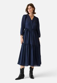 Marineblaues, langärmliges Wickelkleid mit V-Ausschnitt und gestuftem Rock, gefertigt aus einem strukturierten Stoff und mit einem Kordelzug in der Taille. Kombiniert mit schwarzen Stiefeln.