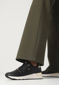 Svarta sportskor med guldkontraster, texturerad yttersko och vit sula. Kombinationen med olivgröna vida byxor visar en avslappnad passform.