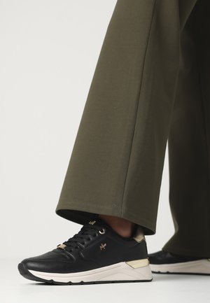 Black athletic shoes with gold accents, textured upper, and a white sole. Paired with olive green wide-leg pants, showing a relaxed fit.