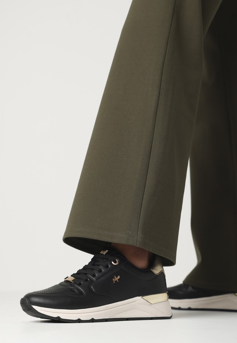 Svarta sportskor med guldkontraster, texturerad yttersko och vit sula. Kombinationen med olivgröna vida byxor visar en avslappnad passform.