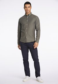 Camisa verde olivo de botones con cuello estructurado, corte ajustado y botones blancos, combinada con pantalones azul marino y zapatillas blancas.