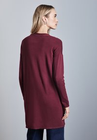Bordeauxfarbener langärmeliger Pullover mit lockerer Passform, Rundhals-Ausschnitt und geripptem Saum. Glatte Textur, getragen über dunkler Jeans.