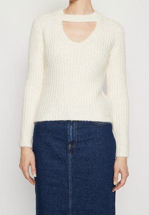 Femme portant un pull en maille côtelée blanc avec une petite découpe au niveau du col à l'avant et une jupe en denim bleu foncé taille haute.
