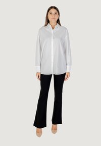 Camicia bianca a maniche lunghe con bottoni e motivo testurizzato, abbinata a pantaloni neri a zampa d'elefante e tacchi nudi, su uno sfondo grigio chiaro.