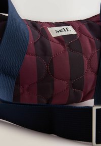 Kvalitatīva somiņa dziļi burgundijas krāsā ar melniem svītrām. Tam ir teksturēta virsma un tumši zilas regulējamas siksnas ar metāla piederumiem.