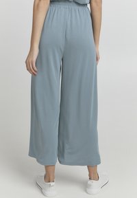 Donna che indossa pantaloni a palazzo larghi e cropped di colore azzurro chiaro e sneakers bianche, mostrata di spalle su uno sfondo semplice.