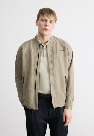 Jaunas vyras dėvi smėlio spalvos BOSS švarką ant šviesios polo marškinėlių ir juodų kelnių, stovintis su rankomis kišenėse prieš baltą foną.