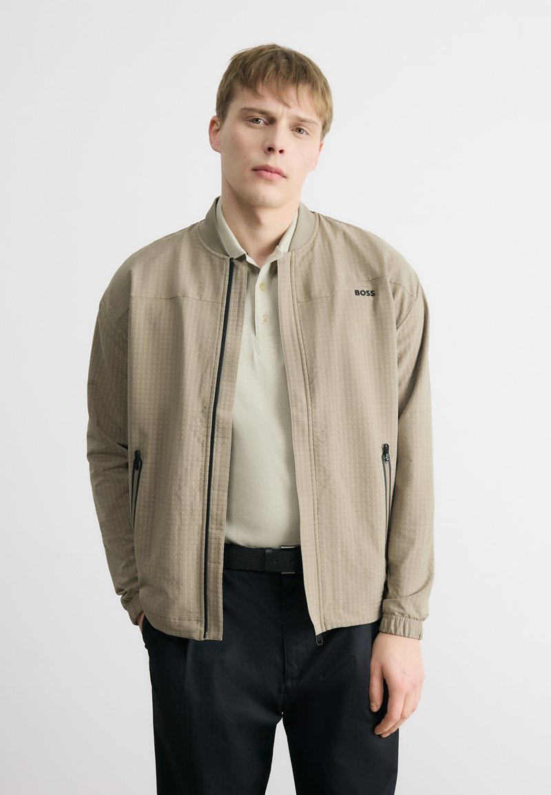 Νεαρός άνδρας που φοράει μπεζ μπουφάν BOSS πάνω από ανοιχτό πολο πουκάμισο και μαύρο παντελόνι, στέκεται με τα χέρια στις τσέπες σε λευκό φόντο.