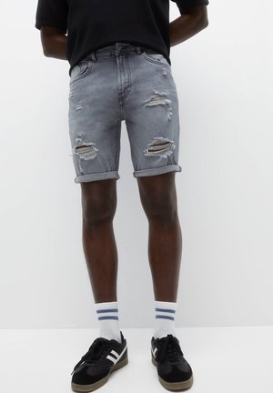Homme portant un short en jean gris déchiré et retroussé, des chaussettes blanches avec des rayures bleues et des baskets noires sur fond blanc.