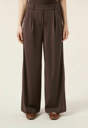 Osoba nosząca luźne, wysokowciskowe szerokie spodnie w ciemnobrązowym kolorze i brązowe buty z zakrytymi palcami, stojąca na tle jednolitego jasnego tła.