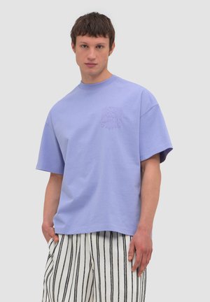 Młody mężczyzna w luźnej lawendowej koszulce z subtelną haftowaną ozdobą na piersi oraz białych spodniach z pionowymi czarnymi paskami, stojący na jednolitym tle.