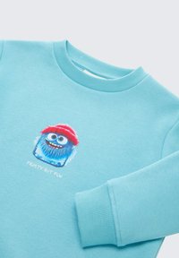 Sweatshirt bleu clair avec col et poignets côtelés. Il présente un graphique de monstre cartoon coloré portant un chapeau rouge et le texte "FROSTY BUT FUN".