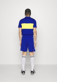 Camiseta deportiva azul con una franja horizontal amarilla, pantalones cortos azules a juego y calcetas blancas hasta la rodilla con rayas negras, llevada con zapatos negros.