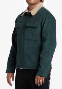 Chaqueta de pana en verde oscuro con un cuello de forro polar beige, que cuenta con un zipper frontal y dos bolsillos en el pecho.