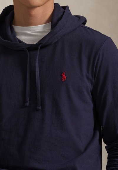 Mees kannab tumerohelist kapuutsiga dressipluusi, millel on nöörid ja punane tikitud polo mängija logo rinnal ning selle all valget ümmarguse kaelusega särk.