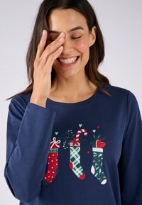 Maglia a maniche lunghe blu navy con tre calze di Natale illustrate in rosso, verde e scozzese, arricchite da elementi decorativi come cuori e bastoncini di zucchero.