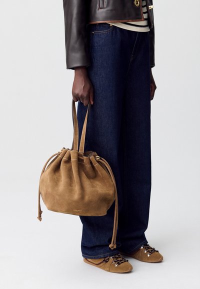 Bolso tipo bucket de ante en marrón claro con cierre tipo ajuste y dos asas. Combinado con jeans de pierna ancha en azul oscuro y zapatos de cordones marrones.