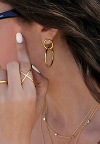 Pendientes de aro dorados con formas circulares y ovaladas entrelazadas. El modelo muestra una mano con múltiples anillos de oro y uñas de un tono pálido.