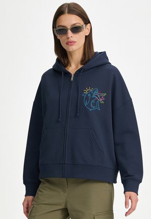 Femme portant un sweat à capuche bleu marine zippé avec un motif requin coloré et un pantalon vert olive, accessoirisée avec des lunettes de soleil rectangulaires foncées.