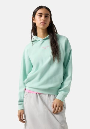 Młoda kobieta z długimi ciemnymi włosami, ubrana w miętową bluzę z kapturem i jasnoszarą spódnicę, stoi na jednolitym tle.