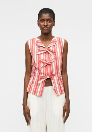 Nő, aki ujjatlan, piros-fehér csíkos felsőt visel három masnival, fehér texturált nadrággal párosítva, egy egyszerű háttér előtt, előre nézve.