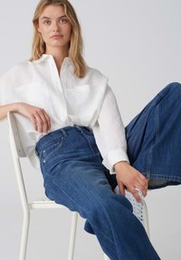 Chemise blanche en lin à boutons avec deux poches avant, associée à un jean large en denim bleu. Mannequin assis sur une chaise blanche, portant des baskets blanches.