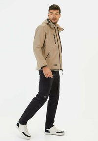 Beige waterdichte jas met een capuchon, rits aan de voorkant en twee zijzakken, gecombineerd met donkere cargobroeken en witte sneakers.