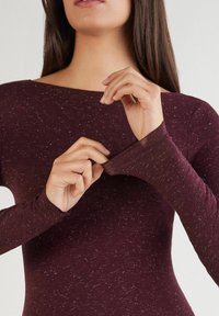 Frau in einem kastanienbraunen, gesprenkelten Langarmshirt, die mit beiden Händen am Ärmelbündchen zieht, vor einem schlichten hellen Hintergrund.