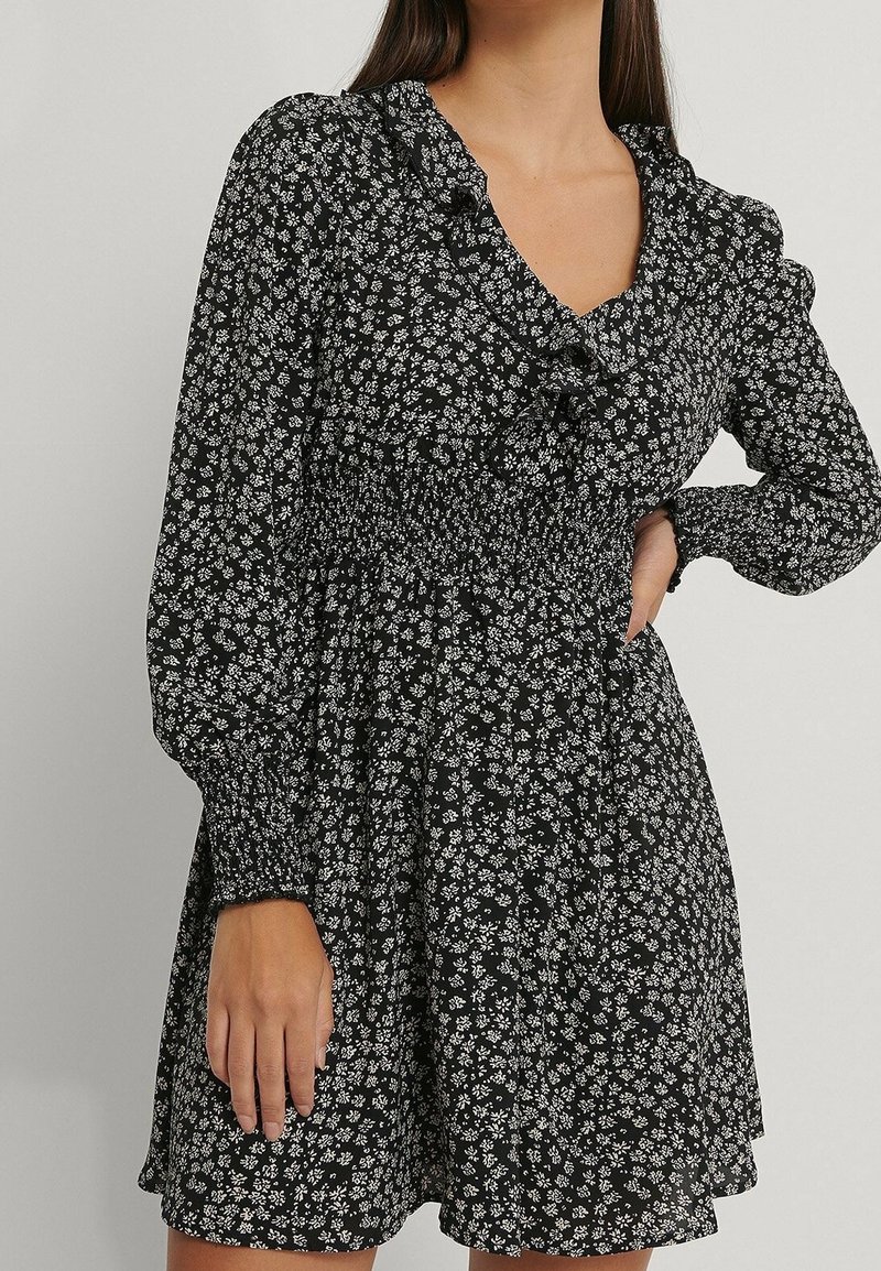 Femme portant une robe noire à manches longues avec un petit motif floral blanc et un col à volants, une main sur la hanche, l'autre reposant le long du corps.