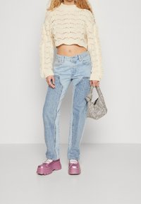 Hellbeige strukturierter, kurzer Pullover mit offener Maschenstruktur. Helle, hoch taillierte Jeans mit kontrastierenden Einsätzen. Dicke, pinkfarbene Plattformschuhe und eine kleine karierte Handtasche.