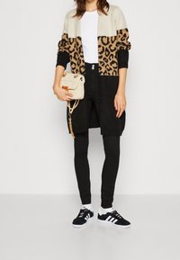 Lange zwarte cardigan met tan en zwart luipaardprint, gedragen over een witte geribbelde top. Gecombineerd met zwarte broeken en zwarte Adidas sneakers.