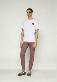White short-sleeve t-shirt with a red circle design, paired with fitted brown pants and dark sneakers with white accents.