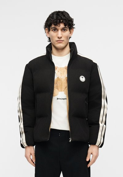 Νέος άνδρας που φοράει μαύρο μπουφάν puffer με λευκές ρίγες στα μανίκια πάνω από λευκό φούτερ με γραφικό αρκουδάκι, στέκεται μπροστά σε απλό φόντο.