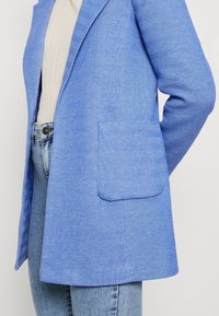 Blazer azul de espiga con un solo bolsillo, forma entallada y textura suave, combinado con jeans azul claros.